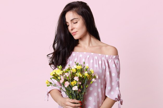 Sideways shot of satisfied pleased European woman with dark hair, holds bouquet of flowers, closes eyes from enjoyment