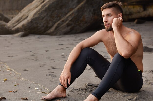 Sideways shot of serious unshaven sportsman sits on sandy beach