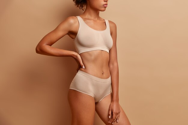 sideways shot of slim woman wears cropped top and panties demonstrates perfect figure after weigh loss poses against beige studio wall