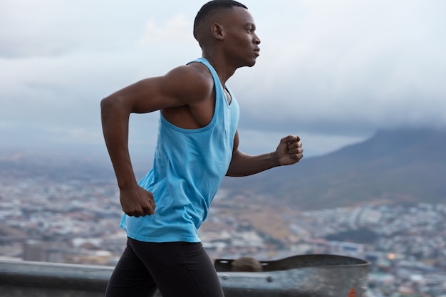 Sideways shot of sporty dark skinned runner wears blue vest, participates in triathlon race, has fitness lifestyle, models against blurred outdoor view with rocks, photographed in fast movement.