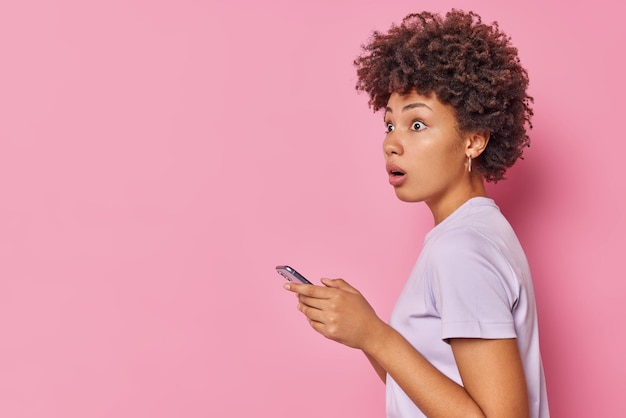 Free photo sideways shot of stunned curly haired woman has shocked concerned expression wears t shirt accidentaly sent message via cellphone wrong person isolated over pink background woth copy space area