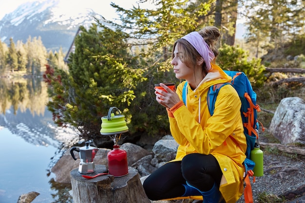 사려 깊은 여행자의 옆쪽 샷은 산 호수 근처의 일회용 컵에서 뜨거운 음료를 즐기고 생각에 깊이 빠져 있습니다.