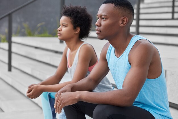 Sideways shot of two black youngsters looks thoughtfully somewhere, pose at stairs, have athletic body, train together, prepare for competition, have thoughtful expressions. Relaxed athletic people