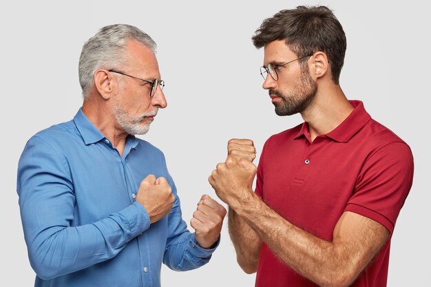 2人の男性の競技者の横向きのショットは、お互いを真剣に見つめ、拳で手を握り締め、戦う準備ができており、共通のビジネスを共有できず、白い壁に立ち向かいます。人と競争