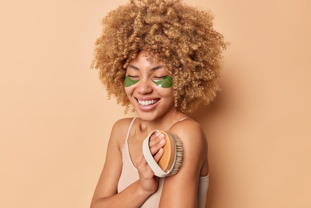 Sideways shot of young woman with curly bushy hair brushes body with wooden brush applies green hydrogel patches under eyes poses against brown studio background. Beauty ad body care concept