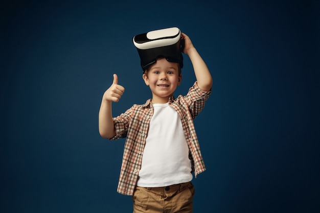 Sign of OK. Little boy or child in jeans and shirt with virtual reality headset glasses isolated on blue studio background. Concept of cutting edge technology, video games, innovation.