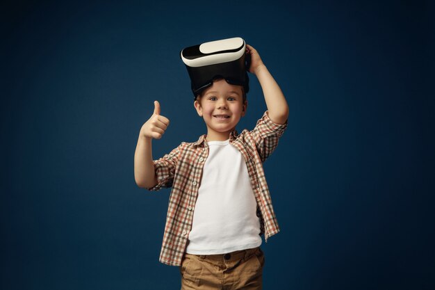 Знак ОК. Маленький мальчик или ребенок в джинсах и рубашке с очками гарнитуры виртуальной реальности, изолированных на синем фоне студии. Концепция передовых технологий, видеоигр, инноваций.