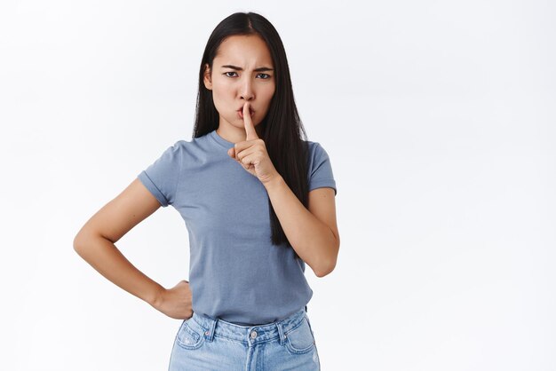 Silence in class Seriouslooking angry bossy asian girl deman quiet say shh press index finger to lips as shushing at you for being loud frowning bothered standing white background