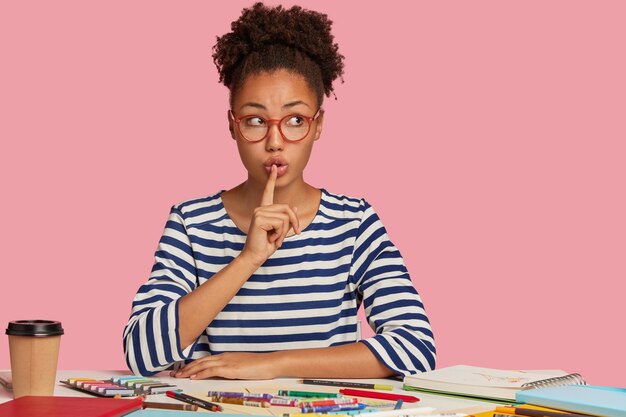 Silent black woman makes shush gesture, has curly hair combed in bun, looks secretly aside, wears striped jumper, asks not make noise, has inspiration for drawing, models over pink  wall