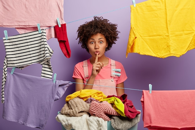 Free photo silent dark skinned housewife hangs clean laundry on clothesline, keeps fore finger over lips, has suprirsed look, busy cleaning clothes, has clothespins on overalls, spreads rumors about neighbour