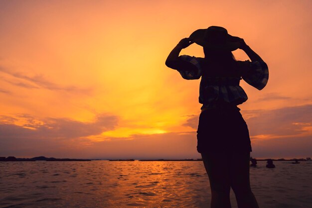 Силуэт азиатской женщины-подростка, поднимающей шляпу к небу на закатном пляже, концепции свободы идей