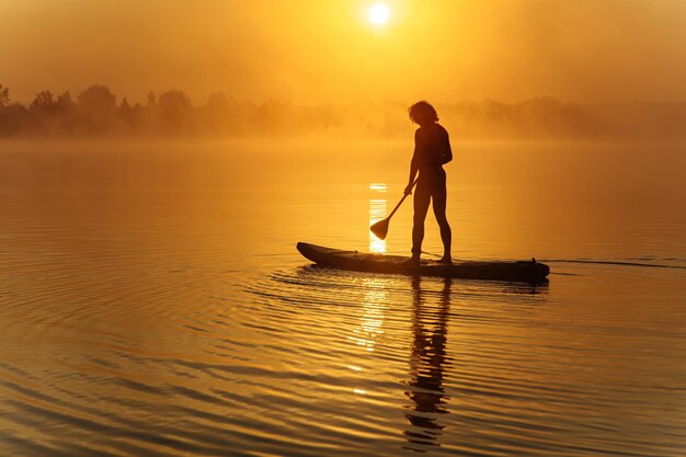 Silhouette of athletic man paddling on board on foggy lake