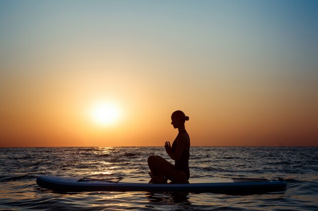 Silhouette of beautiful woman practicing yoga on surfboard at sunrise.