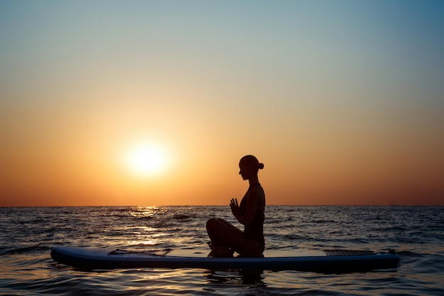 Силуэт йоги красивой женщины практикуя на доске для серфинга на восходе солнца.