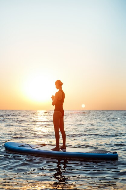 Silhouette of beautiful woman practicing yoga on surfboard at sunrise.