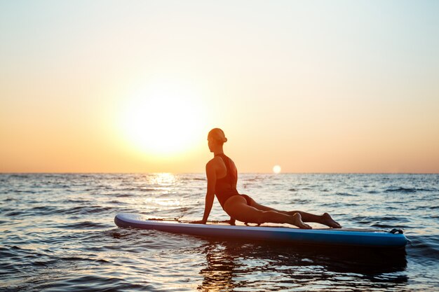 Silhouette of beautiful woman practicing yoga on surfboard at sunrise.