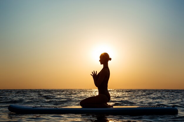 Silhouette of beautiful woman practicing yoga on surfboard at sunrise.