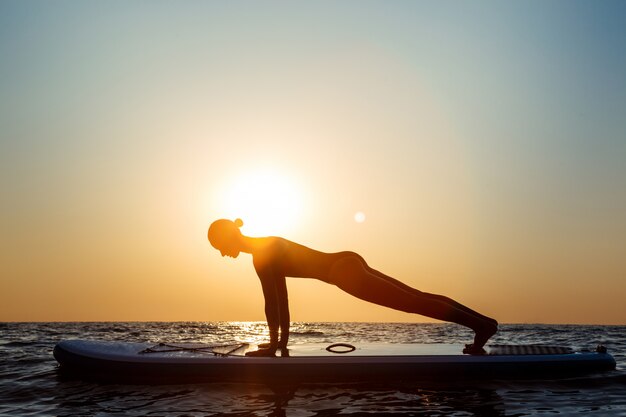 Silhouette of beautiful woman practicing yoga on surfboard at sunrise.