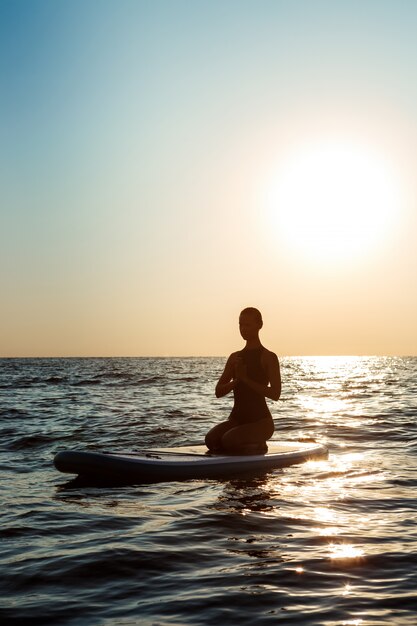 Silhouette of beautiful woman practicing yoga on surfboard at sunrise.