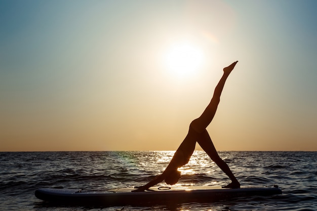 Silhouette of beautiful woman practicing yoga on surfboard at sunrise.