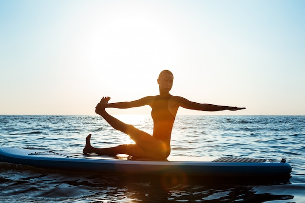 Silhouette of beautiful woman practicing yoga on surfboard at sunrise.