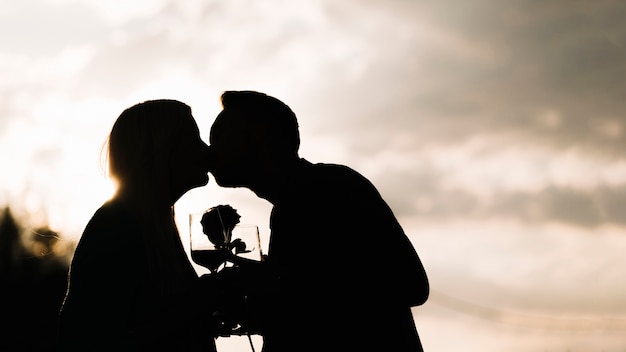 Силуэт пара, проведение бокал и розы, поцелуи против неба