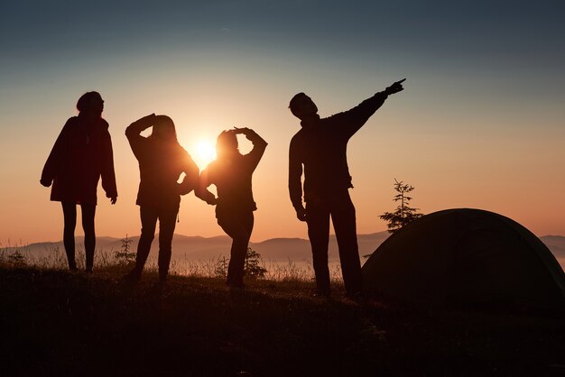 그룹 사람들의 실루엣은 일몰 동안 텐트 근처의 산 꼭대기에서 즐겁습니다.