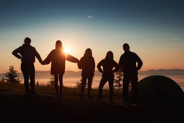 그룹 사람들의 실루엣은 일몰 동안 텐트 근처의 산 꼭대기에서 즐겁습니다.