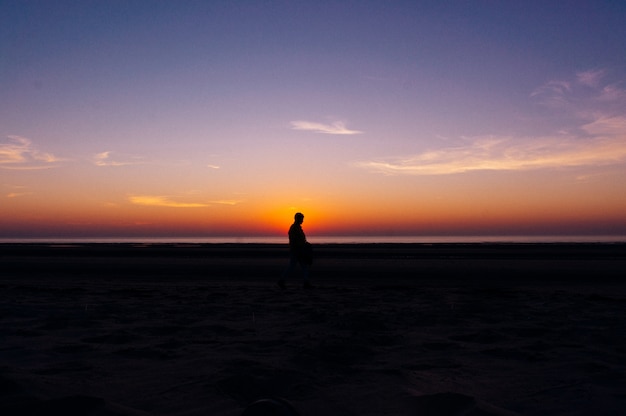 バックグラウンドで夕日の美しい景色とビーチの上を歩く孤独な人のシルエット