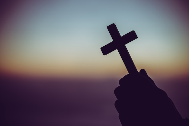 Silhouette of a man praying with a cross in hand at sunrise.