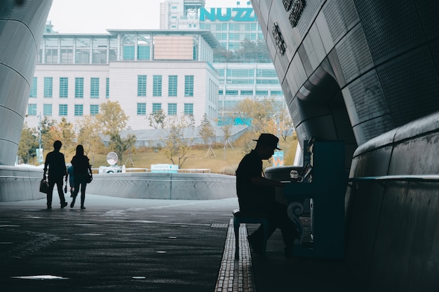 Silhouette of a person in a hat playing the piano outdoors and people walking by