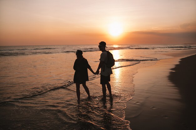 ビーチの上を歩くロマンチックなカップルのシルエットの肖像画。少女と彼女のボーイフレンドが黄金の色鮮やかな夕焼けでポーズ