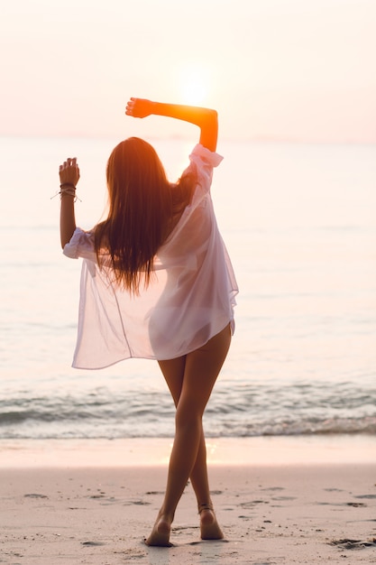 Silhouette of a slim girl standing on a beach with setting sun. She wears white shirt. She has long hair that flies in the air. Her arms stretched into the air