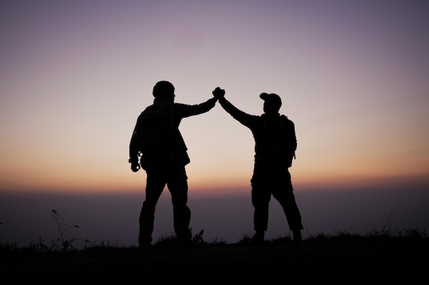 手の信頼を助けるチームワークのシルエット山での成功ハイカーは手を上げて祝う山と夕日の風景の上で互いに助け合う