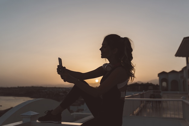 Silhouette of young attractive woman in sportswear making selfie on seafront on sunset. Expressing positivity, active lifestyle, enjoying, cheerful mood.