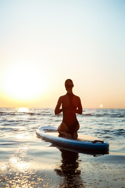 Silhouette of young beautiful girl practicing yoga on surfboard in sea at sunrise.