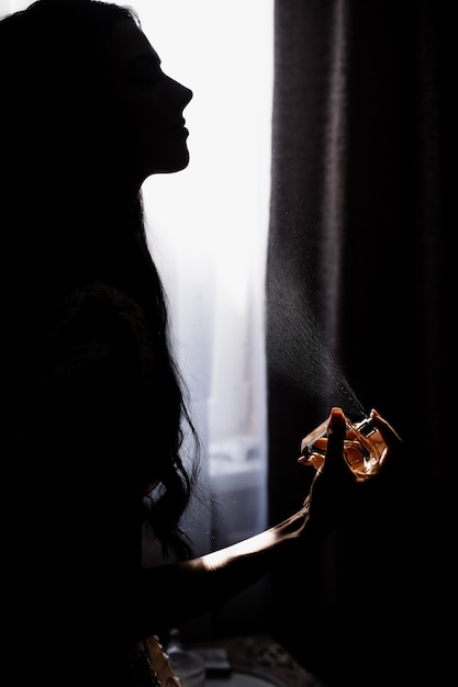 Free photo silhouette of young girl who is perfuming in front of window in darkness