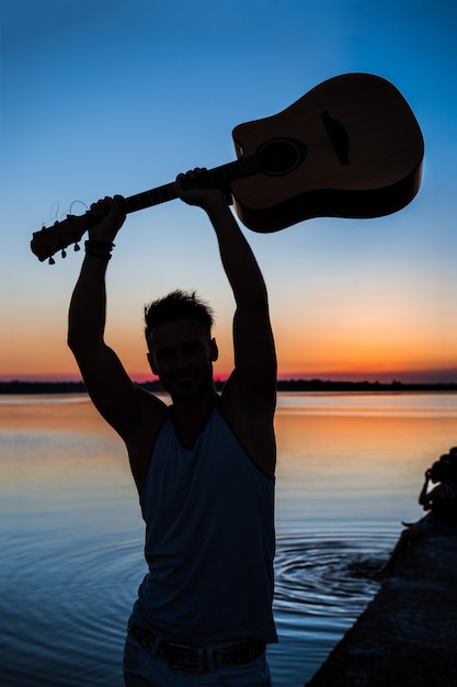 Силуэт молодой красавец, держа гитару на берегу моря во время восхода солнца