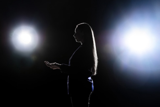 Silhouette of young woman gesturing isolated on black wall with flashlights. Copyspace.