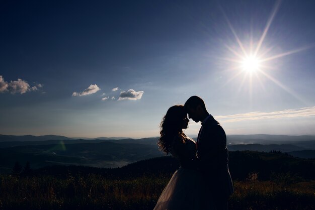 山の風景の前に太陽の光の中に立っている結婚式のカップルのシルエット