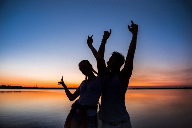 Силуэты молодая красивая пара отдыхает, радуясь на рассвете возле озера