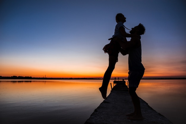 Silhouettes of young beautiful couple resting rejoicing at sunrise near lake