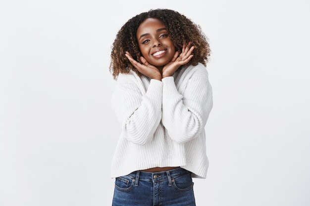 Free photo silly carefree sweet tender african-american curly-haired woman wearing white sweater sighing romantically place hands under jawline smiling delighted passionately look camera daydreaming flirty