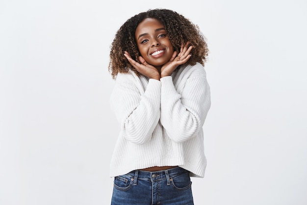 Silly carefree sweet tender african-american curly-haired woman wearing white sweater sighing romantically place hands under jawline smiling delighted passionately look camera daydreaming flirty