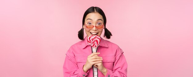Глупая и симпатичная азиатская модель лижет леденец на палочке, ест конфеты сладко и улыбается, выглядя взволнованной