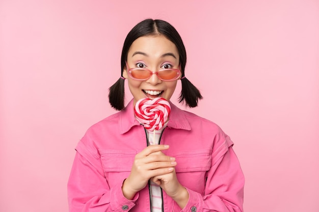 愚かでかわいいアジアの女性モデルは、ピンクの背景の上に立って興奮しているように見える甘いキャンディーを食べてロリポップをなめる