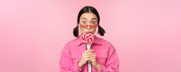 어리 석고 귀여운 아시아 여성 모델이 사탕을 먹고 분홍색 배경 위에 서서 신나게 웃고 있는 롤리팝을 핥습니다.