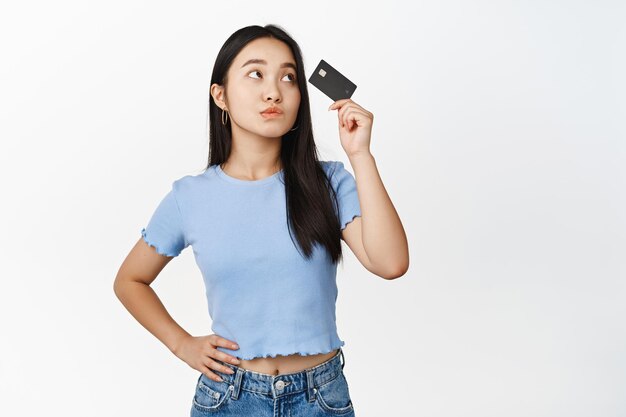 Глупая и милая азиатская девушка смотрит на кредитную карту и думает, стоя задумчиво на белом фоне