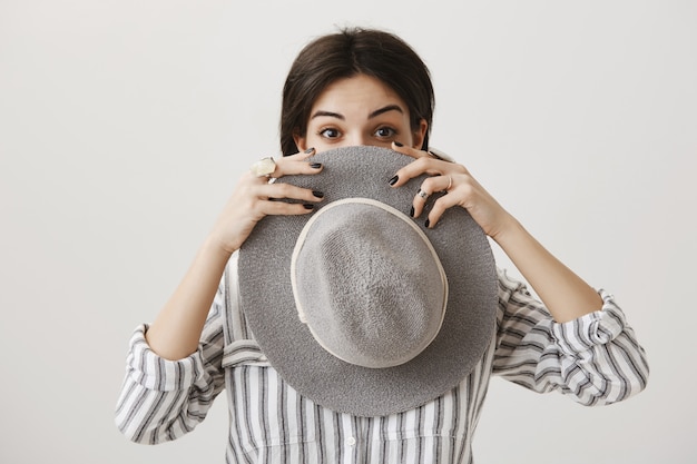 Silly cute brunette female hiding face behind hat and peeking