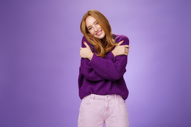 Глупая и милая беззаботная рыжая женщина лет 20, опирающаяся на плечо, обнимая себя, чувствуя тепло в фиолетовом свитере, широко улыбаясь в уютной и расслабляющей атмосфере над фиолетовой стеной.